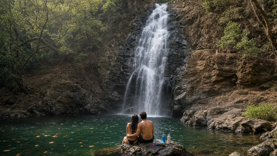 A couple seen from behind sits on a rock by the pool of a tall waterfall falling over dark cliffs